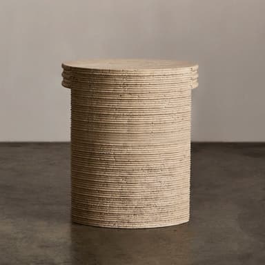 A cylindrical end table with a textured, stone-like surface. The table has a rounded top and a single, curved leg or base element visible. The side of the table exhibits a series of horizontal grooves or ridges, suggesting a layered construction or decorative carving. The material appears porous and natural, consistent with travertine or a similar stone composite. The color is a light, earthy beige. — 3D model