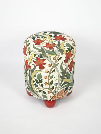 A compact, cylindrical ottoman upholstered in a multi-colored fabric featuring an intricate Arts and Crafts-inspired floral and botanical pattern. The main body of the ottoman rests on four short, spherical, brightly lacquered red feet, providing a contrasting base. The fabric design includes motifs of red flowers, green leaves, and delicate tendrils on an off-white background. — 3D model