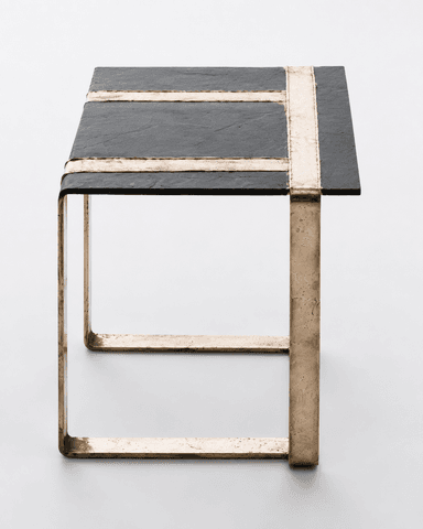 A square side table featuring a dark, textured slate top and a robust, open-cube base constructed from raw, cast bronze. The slate tabletop is inset with two perpendicular strips of the same bronze material, creating a distinctive cross pattern. The bronze frame exhibits a deliberately unfinished, organic texture, emphasizing its handcrafted nature and solid construction. The overall aesthetic is heavy, raw, and industrial, typical of Brutalist design influences. — 3D model