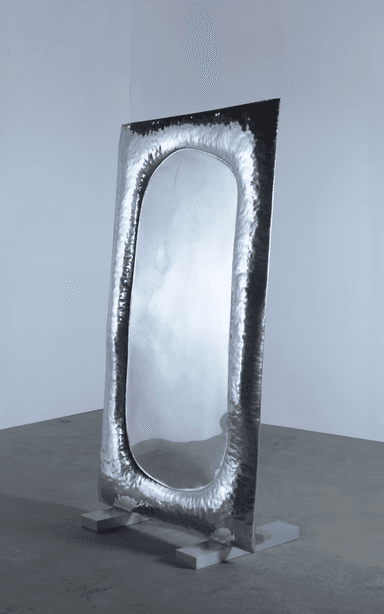 A tall, free-standing mirror with an oval reflective surface. The frame surrounding the mirror is irregular and appears to be made of hammered metal, giving it a textured, artisanal look. The mirror is supported by two rectangular grey bases. — 3D model