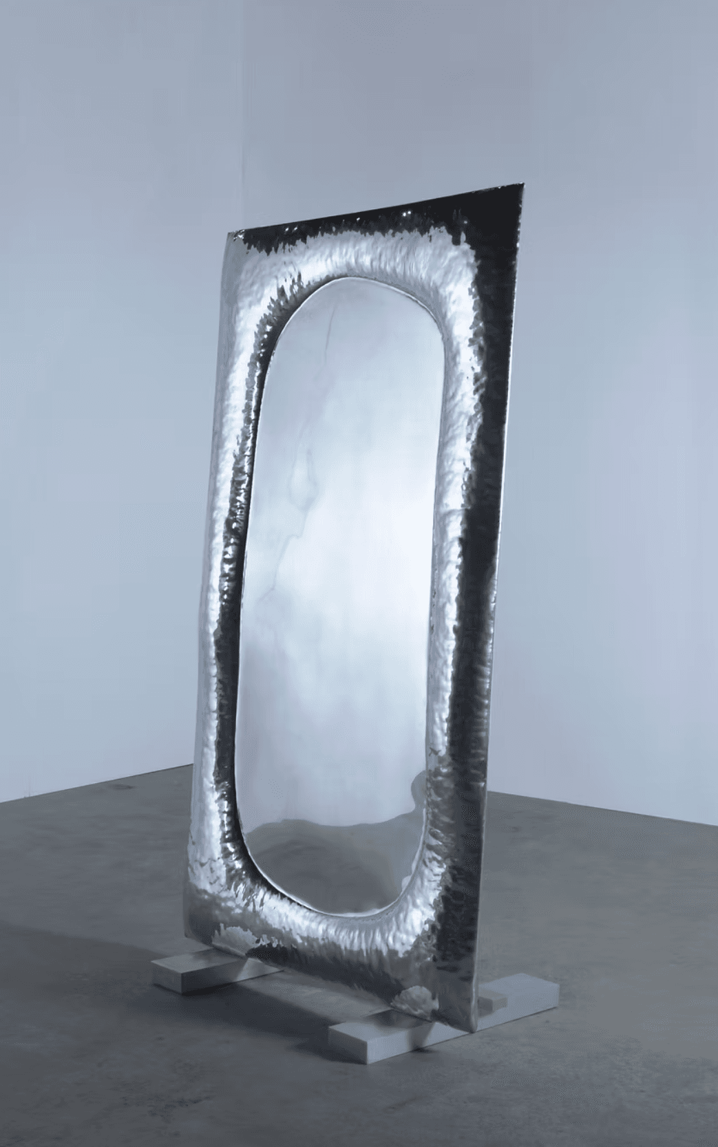 A tall, free-standing mirror with an oval reflective surface. The frame surrounding the mirror is irregular and appears to be made of hammered metal, giving it a textured, artisanal look. The mirror is supported by two rectangular grey bases. — 3D model