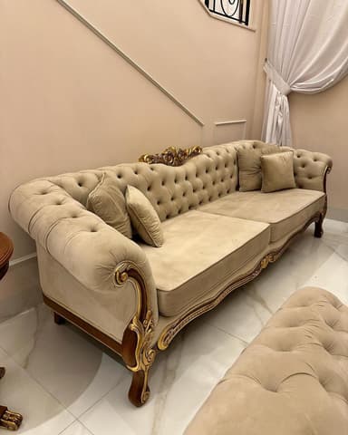 A luxurious, large sofa featuring a deep button-tufted backrest and roll arms, upholstered in a light beige velvet or similar plush fabric. The sofa boasts an intricately carved wooden frame with extensive gilded detailing, particularly visible along the base, armrests, and upper back. It includes two seat cushions and several matching throw pillows. The overall design evokes an opulent, classical, or baroque aesthetic. — 3D model