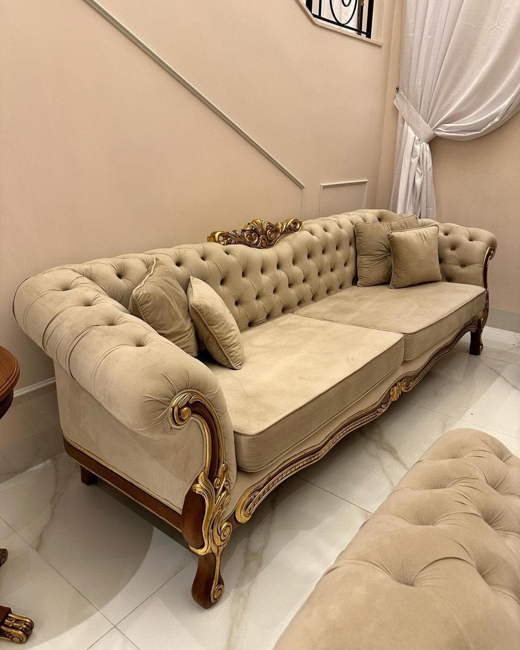 A luxurious, large sofa featuring a deep button-tufted backrest and roll arms, upholstered in a light beige velvet or similar plush fabric. The sofa boasts an intricately carved wooden frame with extensive gilded detailing, particularly visible along the base, armrests, and upper back. It includes two seat cushions and several matching throw pillows. The overall design evokes an opulent, classical, or baroque aesthetic. — 3D model