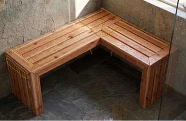 An L-shaped bench constructed from light-toned wooden slats, featuring a contemporary design. The slatted surfaces are present on both the seat and the solid block-style legs, creating a cohesive and sturdy appearance. The bench is designed to fit into a corner, providing versatile seating. — 3D model