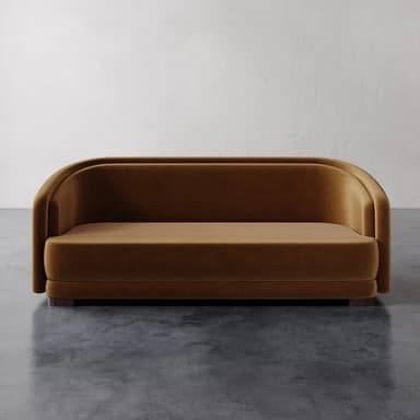 A contemporary loveseat featuring a continuous, rounded backrest and integrated arms upholstered in a plush, camel-colored velvet fabric. The design is characterized by its smooth, curvilinear lines and minimalist profile, resting on a recessed dark wood base. The seat cushion is integrated, contributing to the streamlined appearance. — 3D model