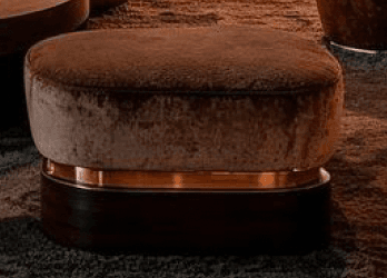 A contemporary ottoman featuring a rounded square form. The top cushion is upholstered in a textured, light brown fabric, resembling velvet or chenille. Below the upholstered top, there is a polished rose gold or copper metallic band, which transitions into a dark, rich-toned wooden base. The wood exhibits a prominent, horizontal grain pattern, contributing to its sophisticated appearance. The overall design suggests a blend of modern elegance and luxurious materials. — 3D model