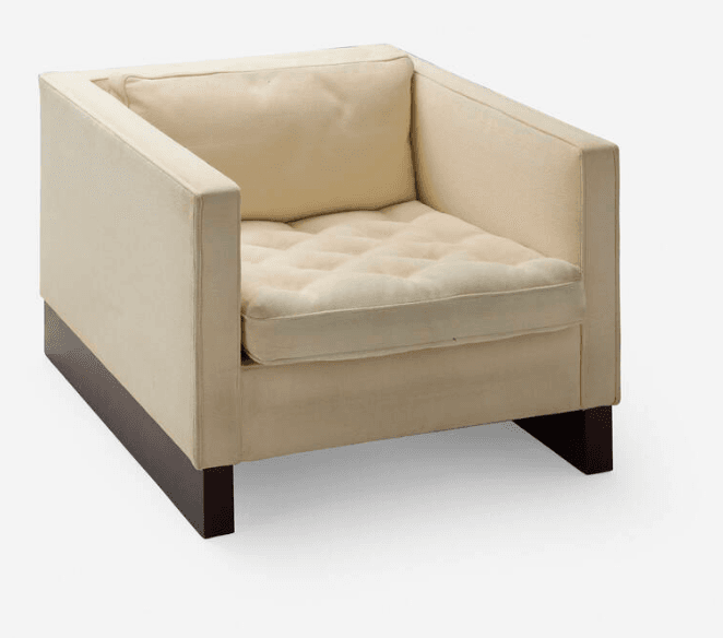 A modern armchair with a blocky, rectilinear design. The chair features a tufted seat cushion and a separate back cushion, both upholstered in a light-colored fabric. The frame is also upholstered in the same fabric, creating a seamless appearance. The armchair is supported by a low, dark-colored base and four short, square legs. — 3D model