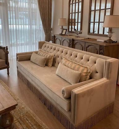 A sophisticated long sofa featuring deep button tufting across its high backrest and inner arm panels. The upholstery is a luxurious champagne beige velvet, complemented by a decorative fringed skirt along the base. The sofa is adorned with three rectangular throw pillows and two bolster pillows, all covered in coordinating or subtly patterned fabric. Its design features straight, high arms that align with the backrest, contributing to its formal and elegant aesthetic. — 3D model