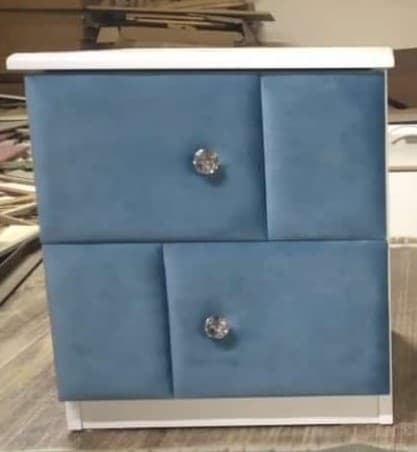 A compact, modern nightstand featuring a clean white frame and two drawers with soft, blue upholstered fronts. Each drawer is adorned with a clear, crystal-like knob, adding a touch of elegance to the piece. — 3D model