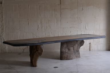 A large dining table with an irregular, organic-shaped top and two substantial, textured bases that resemble natural rock formations or tree trunks. The tabletop appears to be made of multiple panels, possibly wood or stone, with a dark, possibly metal or resin, edge banding. The bases are dark brown and have a rough, sculpted texture, giving the impression of raw, natural elements. — 3D model