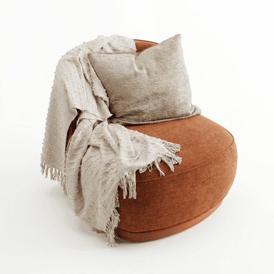 A contemporary armchair with a rounded silhouette, upholstered in a textured, rust-colored fabric. It features a low profile with a visible wooden base. A textured throw blanket with fringes and a decorative pillow are draped over the back and armrest. — 3D model