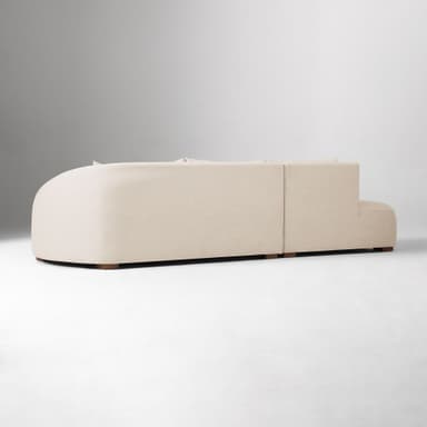 A sectional sofa with a chaise lounge. The sofa has a low profile and is upholstered in a textured fabric. It features plush back cushions and a rounded armrest on the chaise end. The sofa is supported by small, dark wooden block feet. — 3D model