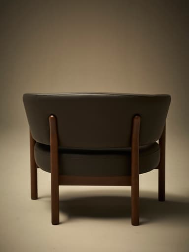 A low-profile armchair with a dark brown leather upholstered seat and curved backrest. The frame and legs are constructed from dark wood, exhibiting a minimalist design. The seat and back appear to be separate components supported by the wooden frame. — 3D model