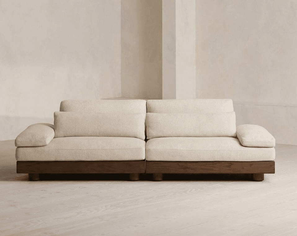 A modern, low-profile sofa with a sectional appearance, featuring a thick, upholstered base and seat cushions. The sofa has distinct armrests and back cushions. It is elevated by a dark wooden plinth base with cylindrical feet. — 3D model