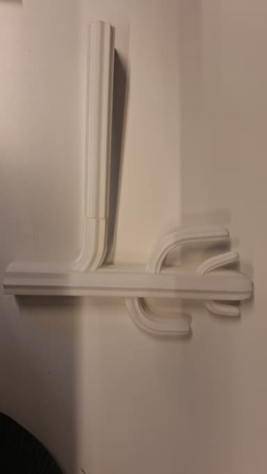 A minimalist, wall-mounted coat rack rendered in a uniform white finish. It features a vertical rectangular mounting post that connects to a horizontal bar, from which multiple curved hooks extend. Both the vertical post and the horizontal bar are adorned with a fluted or paneled design, giving the object a sculptural and architectural quality. The hooks are smoothly curved and rounded, designed to hold garments or accessories. — 3D model