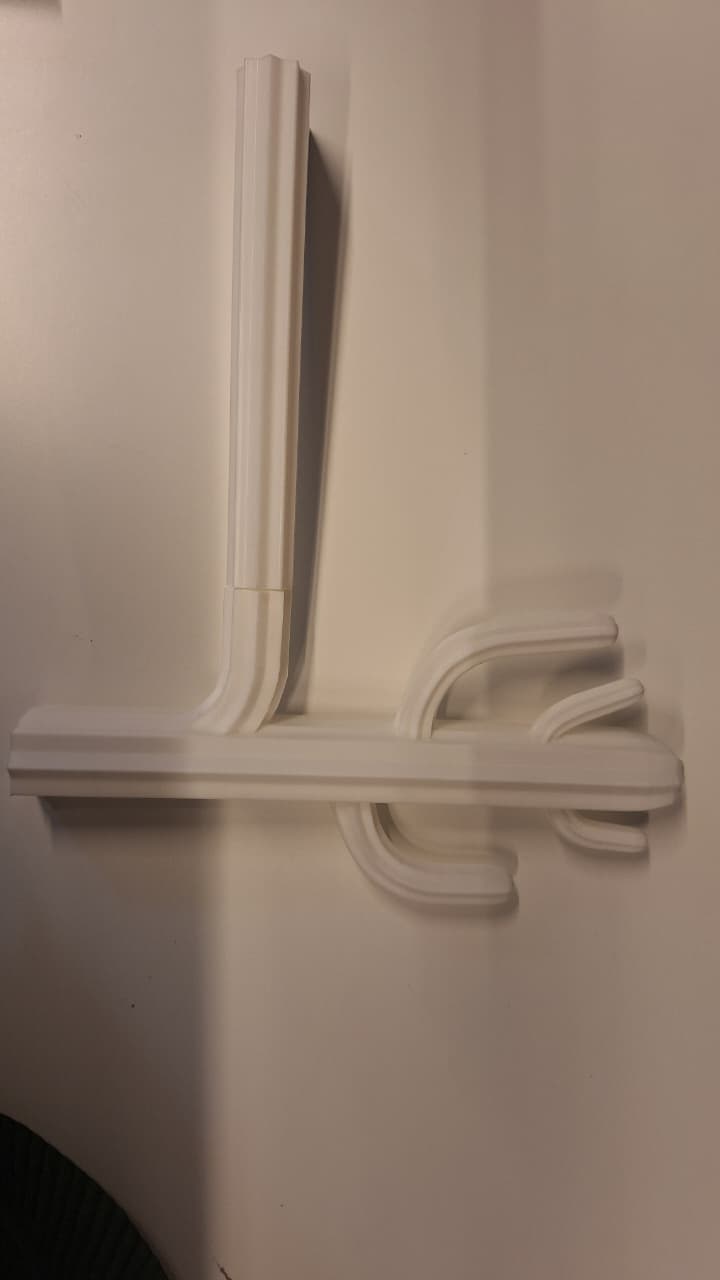 A minimalist, wall-mounted coat rack rendered in a uniform white finish. It features a vertical rectangular mounting post that connects to a horizontal bar, from which multiple curved hooks extend. Both the vertical post and the horizontal bar are adorned with a fluted or paneled design, giving the object a sculptural and architectural quality. The hooks are smoothly curved and rounded, designed to hold garments or accessories. — 3D model
