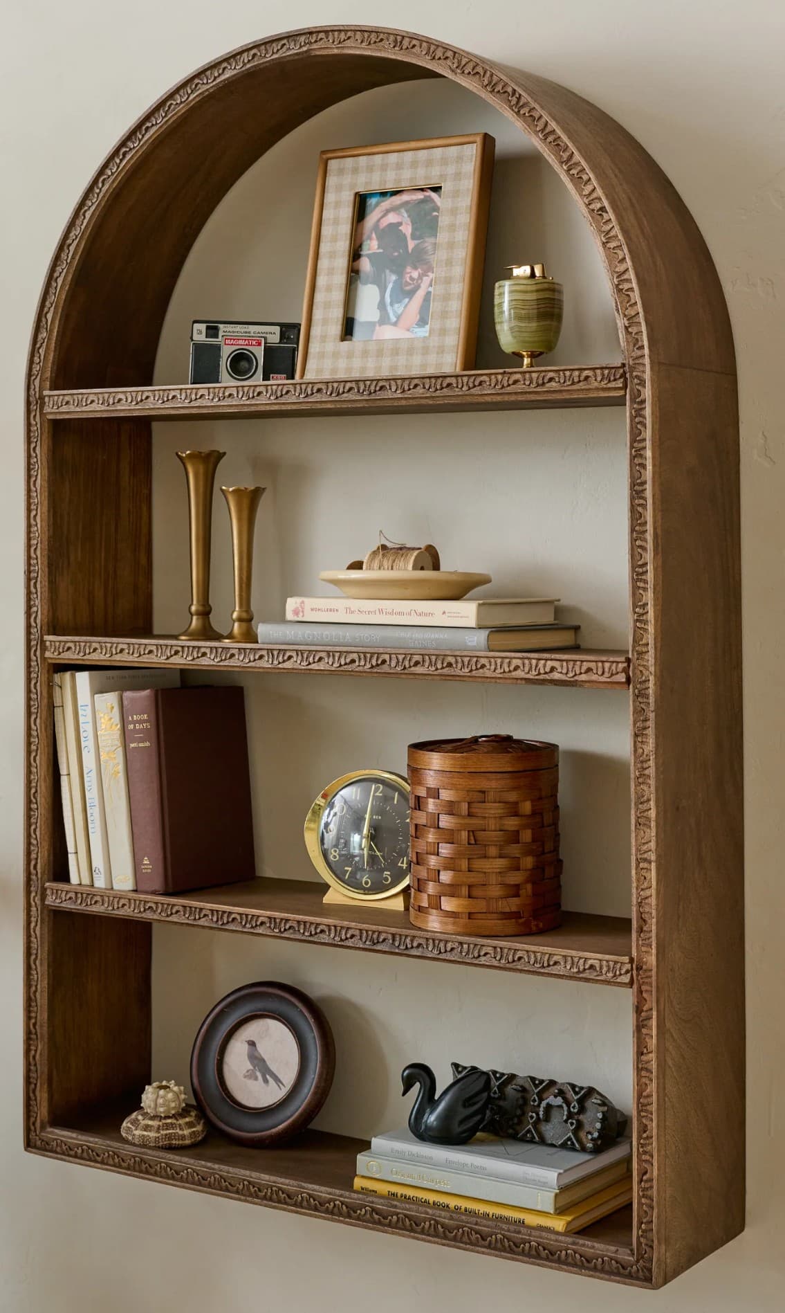 A wooden bookshelf with an arched top and three shelves. The frame and edges of the shelves are adorned with carved decorative motifs. The top shelf displays a framed photograph, a camera, and a small decorative vase. The second shelf holds two vases, a shallow dish with decorative items, and two books. The third shelf contains several books, a round analog clock on a stand, and a woven basket. The bottom shelf features a framed picture, decorative objects including a small sculpture of a swan and an ornamental piece, and a stack of books. — 3D model