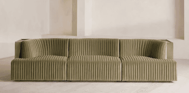 A long, contemporary sofa with a channeled upholstery design. The sofa features a modular appearance with distinct sections, creating a sense of depth and texture. It has a low profile and a deep seat, with curved armrests or side panels. A small, dark wood-toned surface is visible on the far left side, integrated into the structure. — 3D model