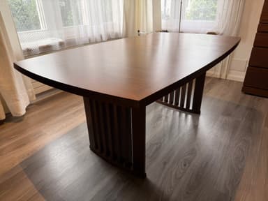A modern dining table featuring a dark brown wooden top with elegantly curved edges and a matching slatted base. The table top displays a visible wood grain pattern, suggesting a veneer finish, and shows some minor surface wear. The base appears robust, designed with vertical wooden slats, which likely form a pedestal or trestle-style support. — 3D model