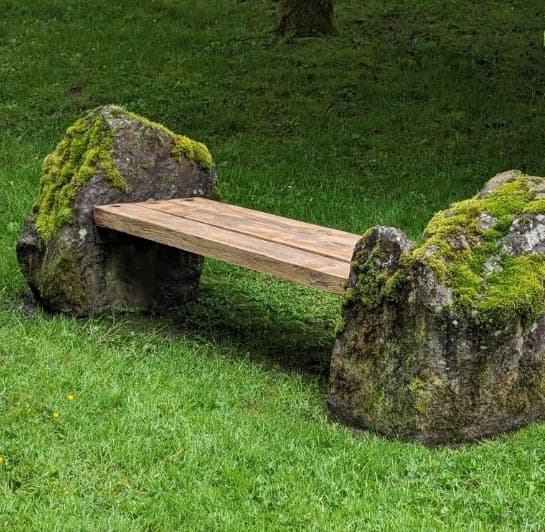 An outdoor bench composed of three wooden planks forming the seat, supported by two large, moss-covered natural stone blocks as legs. — 3D model