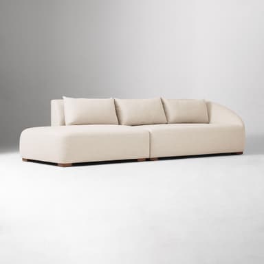 A sectional sofa with a chaise lounge. The sofa has a low profile and is upholstered in a textured fabric. It features plush back cushions and a rounded armrest on the chaise end. The sofa is supported by small, dark wooden block feet. — 3D model