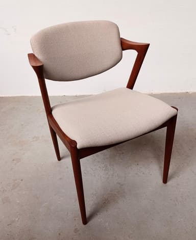 A mid-century modern armchair with a teak wood frame and upholstered seat and backrest. The frame features a distinctive angular design with tapered legs and elegantly curved armrests. The upholstery is a textured, light-colored fabric. — 3D model