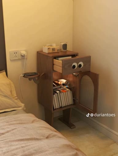 A whimsical wooden nightstand featuring a single drawer with decorative "googly eyes" and a lower cabinet with a reeded glass door. An integrated round shelf extends from the side, designed to hold a small device like a smartphone. The unit stands on two broad, flat feet. — 3D model