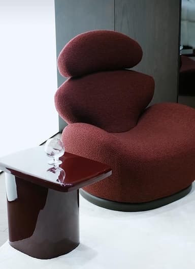 A postmodern sculptural lounge chair, upholstered in a deep burgundy, textured fabric. The design features multiple organic, rounded forms stacked vertically, creating a fluid and continuous shape without distinct arms or backrest separations. It rests on a dark, circular, flat base. — 3D model