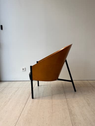 A striking mid-century modern lounge chair featuring a gracefully curved, light-brown plywood shell that forms both the backrest and integrated armrests. The chair is supported by a minimalist black metal frame with slender legs. A dark, possibly black or dark brown, upholstered cushion rests on the seat, contrasting with the warm wood tones. — 3D model