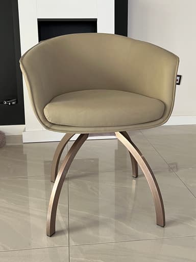 A modern accent chair with a curved, upholstered back and seat. The chair is supported by a distinctive base composed of four curved legs that splay outwards, made of a metallic material. A vertical seam with a zipper-like detail runs up the center of the chair's back. — 3D model