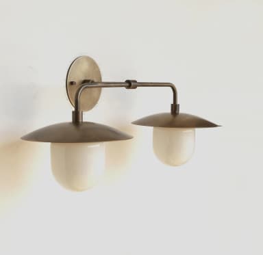 A wall-mounted lighting fixture featuring two light sources, constructed from aged brass or bronze metal, with each light housed under a conical metal shade and diffusing light through an opaque, white, bell-shaped glass component. — 3D model