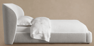 A close-up view of a modern upholstered bed frame, featuring a softly curved, enveloping headboard and side rails covered in a textured, light-colored boucle fabric. A white pillow and bedsheet are visible, indicating its function. — 3D model