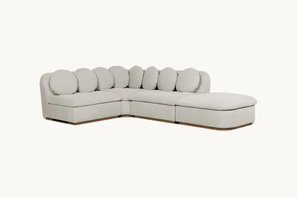 A modular sectional sofa with a chaise lounge. The sofa features a low profile with square seat cushions and rounded back cushions arranged in a series. It has a light-colored fabric upholstery and a visible wooden base. — 3D model
