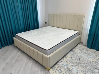 A modern platform bed with a tall, slightly angled headboard and a matching low-profile base, both upholstered in a light beige fabric. The headboard and bed frame feature distinctive vertical channel tufting. Small, possibly metallic, feet are visible at the corners, and a white mattress with a dark border rests on the platform. — 3D model