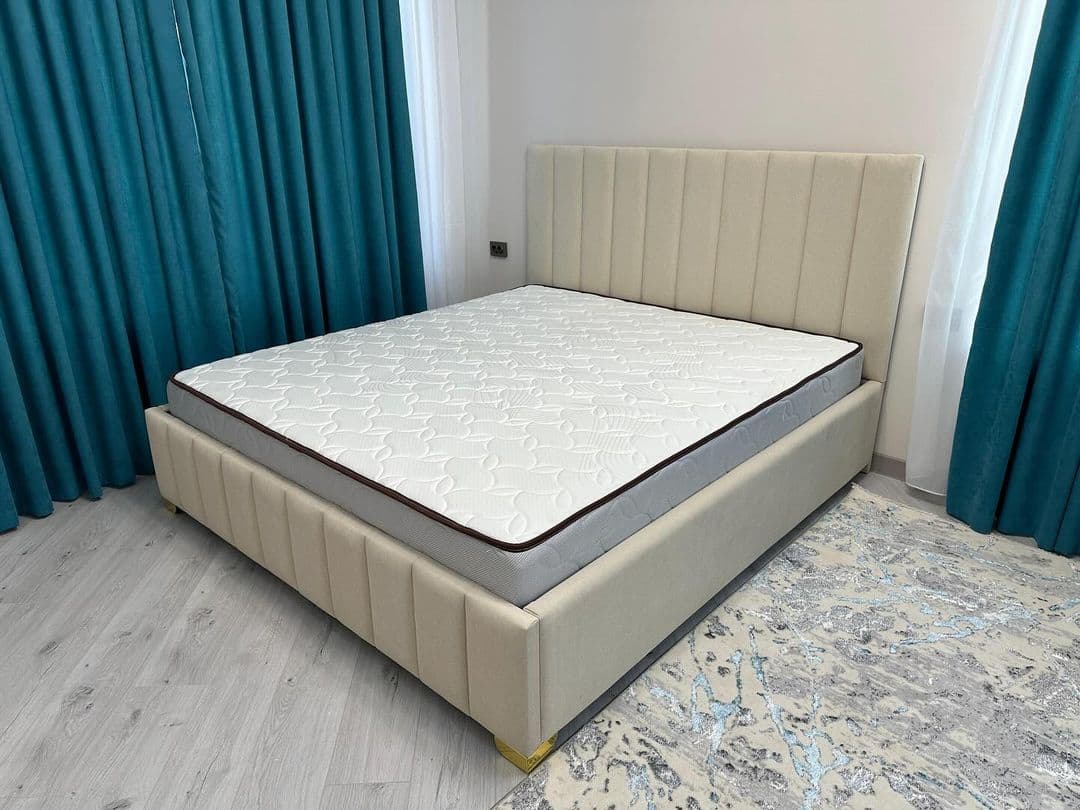 A modern platform bed with a tall, slightly angled headboard and a matching low-profile base, both upholstered in a light beige fabric. The headboard and bed frame feature distinctive vertical channel tufting. Small, possibly metallic, feet are visible at the corners, and a white mattress with a dark border rests on the platform. — 3D model