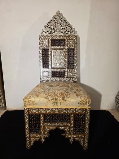 A highly ornate accent chair featuring intricate marquetry and inlay work. The chair has a tall, elaborately carved back with floral motifs and geometric patterns. The seat is upholstered in a light-colored fabric with a floral pattern. The base of the chair, including the legs and the area beneath the seat, is constructed from dark wood and heavily decorated with mother-of-pearl inlay, creating floral and geometric designs. The legs have an arched structure. The overall construction suggests a piece of antique or artisan craftsmanship. — 3D model