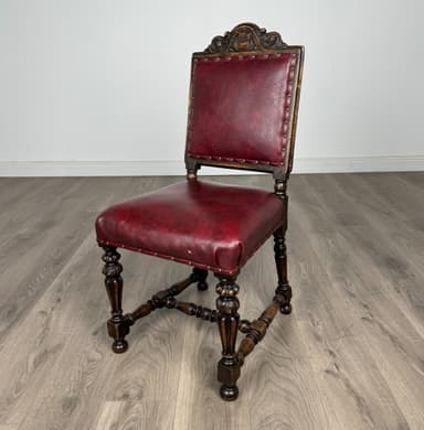 An ornate, traditional side chair featuring a dark carved wood frame and burgundy leather upholstery. The backrest has an elaborate carved crest rail and stiles, while the seat and back are padded and trimmed with individual brass nailheads. The legs are turned and carved, connected by stretchers, and terminate in bun feet. — 3D model