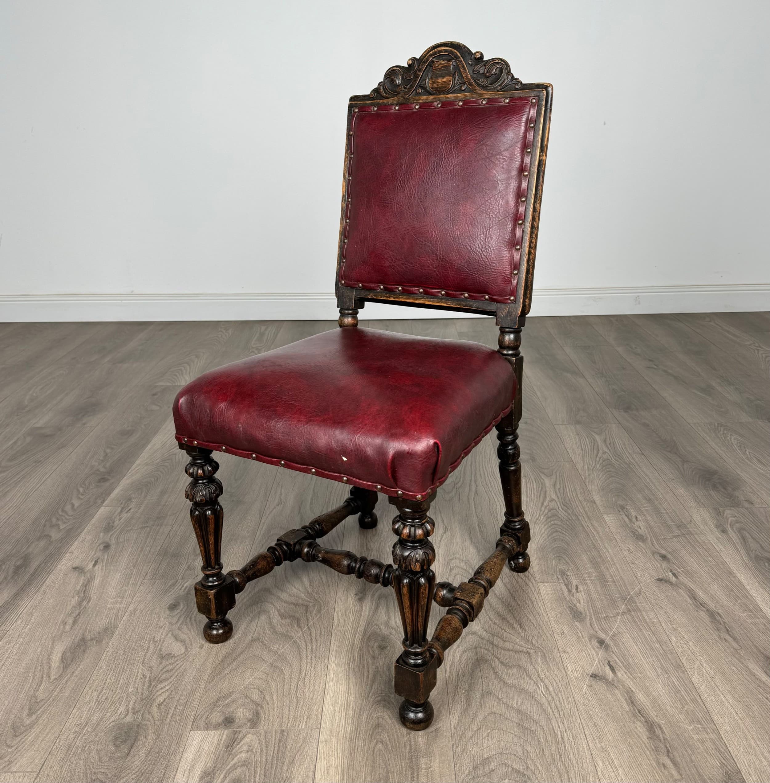 An ornate, traditional side chair featuring a dark carved wood frame and burgundy leather upholstery. The backrest has an elaborate carved crest rail and stiles, while the seat and back are padded and trimmed with individual brass nailheads. The legs are turned and carved, connected by stretchers, and terminate in bun feet. — 3D model