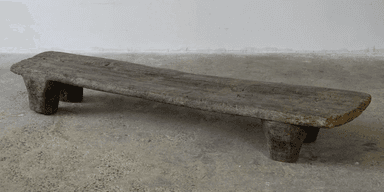 A rustic, low-profile bench with a flat, elongated top and two substantial, block-like legs at each end. The piece exhibits a weathered texture and irregular edges, suggesting it is made from natural wood. The design is simple and primitive. — 3D model