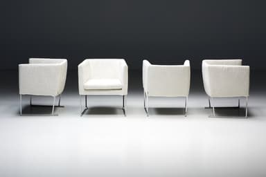 A set of four identical armchairs with a modern design. Each armchair features a cube-like upholstered seat and backrest with squared arms. The upholstery appears to be a textured fabric. The chairs are supported by a sleek, minimalist frame, likely made of polished metal, elevating them slightly off the ground. The overall aesthetic suggests a contemporary or minimalist style. — 3D model