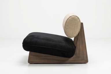 A modern, low-profile lounge chair featuring a distinctive solid wood base. The base is dark-toned wood, shaped with a geometric, wedge-like profile. It supports a plush, rectangular black seat cushion, likely upholstered in velvet or microsuede. The backrest is a cylindrical bolster cushion, upholstered in a textured light beige fabric, resting against the angled wooden support structure. — 3D model