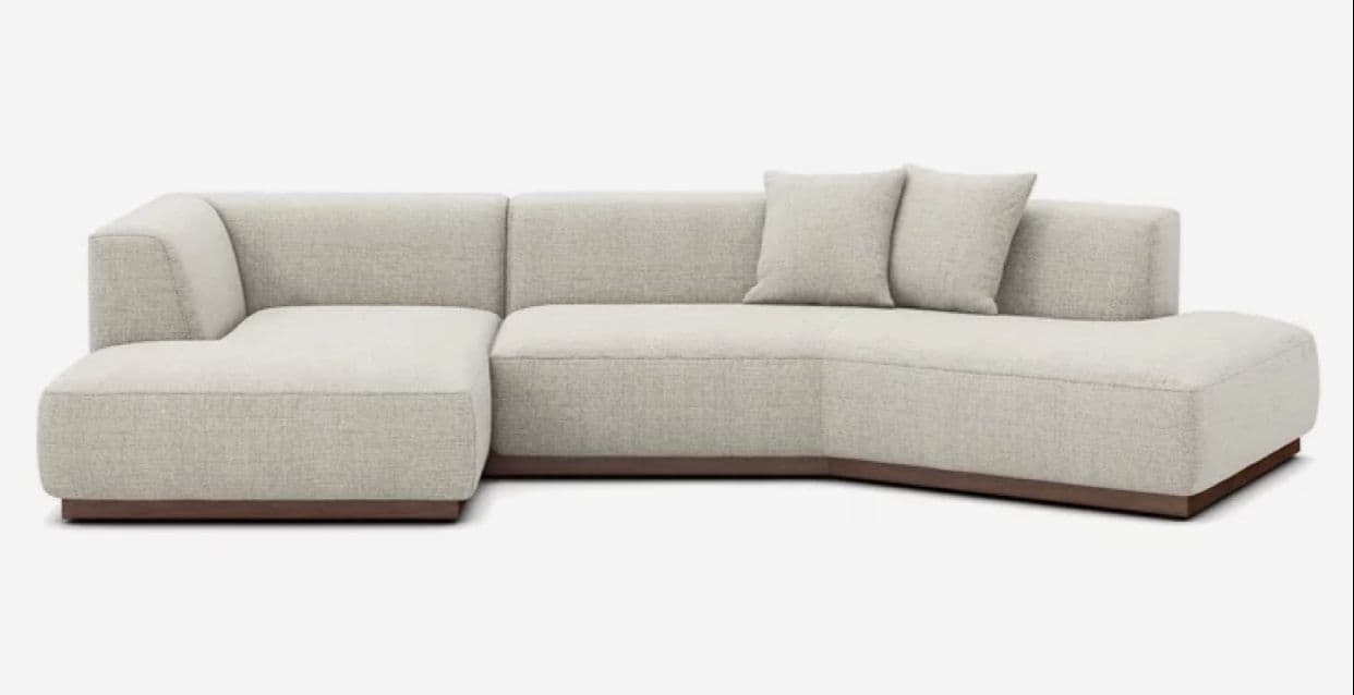 A modern, L-shaped sectional sofa upholstered in a light, textured fabric, featuring a modular design with an integrated chaise lounge on one end and an armless section. It sits on a recessed, dark wood plinth base. Two matching throw pillows are included. — 3D model