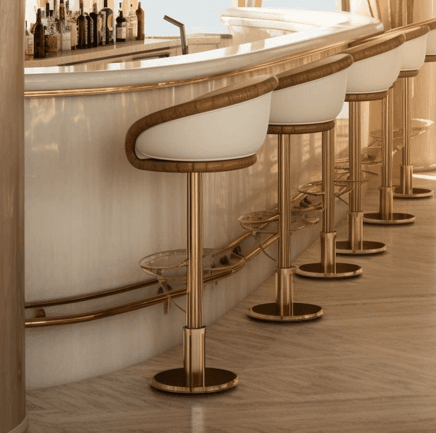 A modern bar stool featuring a barrel-style seat upholstered in white faux leather with a tan leatherette trim. It has a gold-finished metal pedestal base with a circular disc base and an integrated semi-circular glass footrest. The seat appears to be padded and includes gold piping detail. — 3D model