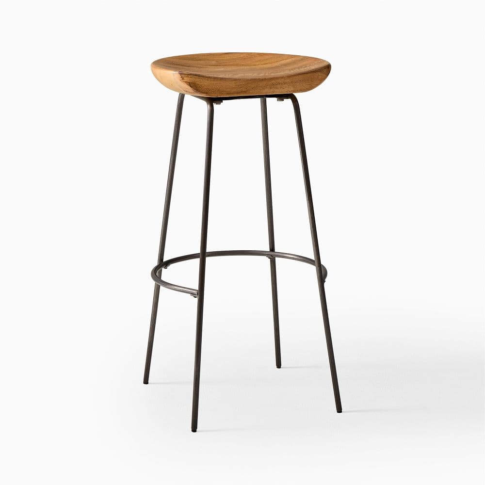 A tall bar stool featuring a sculpted, saddle-shaped wooden seat and a minimalist metal rod base with an integrated footrest. The wood seat has a visible grain and a warm, natural finish, while the metal legs are slender and finished in a dark, possibly black or dark gray, color. The four legs splay slightly for stability and are connected by a circular ring that serves as a footrest. The design is clean and functional, combining natural and industrial elements. — 3D model