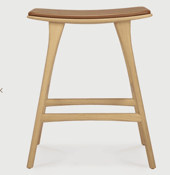 A modern bar stool featuring a solid light-colored wood frame and a contoured, saddle-shaped seat upholstered in tan leather. The stool has four tapered legs connected by two horizontal stretchers acting as footrests. Its design is minimalist and ergonomic, emphasizing natural materials and clean lines. — 3D model