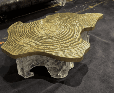 A striking coffee table featuring an irregular, organic-shaped tabletop made of polished golden brass, adorned with a detailed concentric ring pattern. The base is crafted from a chunky, translucent material, likely clear resin or acrylic, sculpted into an abstract, ice-like form, creating a visually compelling contrast with the metallic top. The table exhibits a low profile. — 3D model