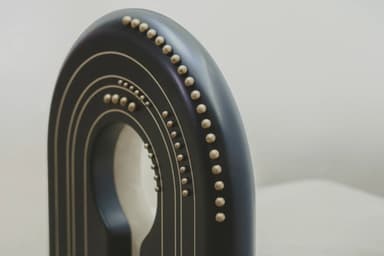 A contemporary chair with a distinctive U-shaped backrest that forms a cutout. The seat and the curved portion of the backrest are upholstered in a light beige, suede-like fabric. The frame is composed of dark, possibly black or dark wood, elements. The outer curved frame of the backrest and the vertical supports of the legs are adorned with a series of small, brass or gold-colored studs. The legs are thick and rectangular, with a dark finish. The chair's design is modern and somewhat sculptural, featuring a negative space in the upper backrest. — 3D model
