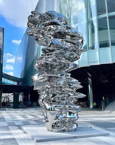A tall, abstract floor sculpture with an organic, twisted form, rendered in a smooth, polished white material, likely resin or fiberglass. It stands on a simple square base, illuminated by uplighting, within an interior space characterized by ancient-looking brick walls. — 3D model