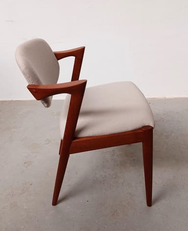 A mid-century modern armchair with a teak wood frame and upholstered seat and backrest. The frame features a distinctive angular design with tapered legs and elegantly curved armrests. The upholstery is a textured, light-colored fabric. — 3D model