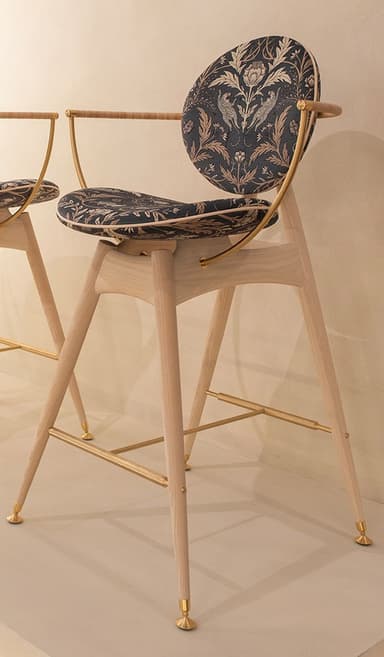 A tall, contemporary bar stool featuring a light-toned wooden frame, brass accents, and distinctive dark, patterned upholstery. The seat and backrest are circular and upholstered with a black fabric featuring an intricate floral and animal pattern in gold and bronze tones. The backrest is supported by curved brass elements that also form integrated armrests. The splayed wooden legs are reinforced by brass stretchers that serve as a footrest and terminate in brass caps. — 3D model